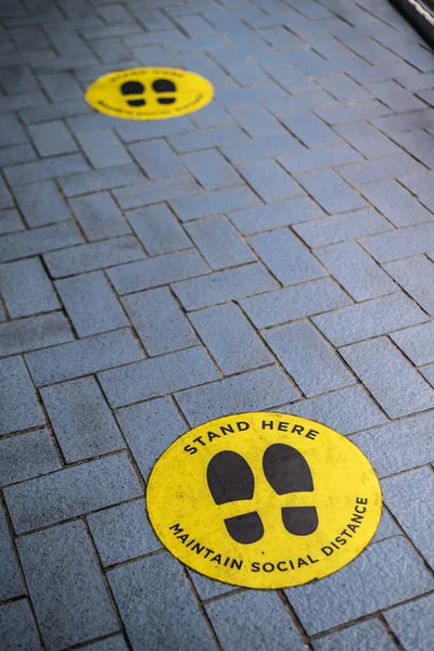 Social distancing sign stickers plastered on a brick floor near the entrance of a mall or shopping plaza. 2 stickers spaced apart at recommended distance.