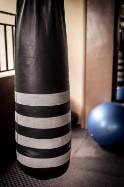 Eski siyah beyaz bir kum torbası. Yanında mavi bir spor topu var. Sert bir spor salonunun içinde..