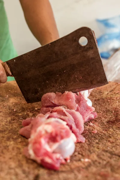 Chopping up a piece of pork shoulder on a large wood chopping block with a large cleaver knife at a kitchen or meat shop.