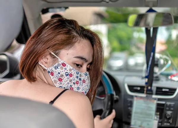 アジア系の女性が車を運転しながら後部座席を見る 布面マスクを着用 自然光撮影 — ストック写真