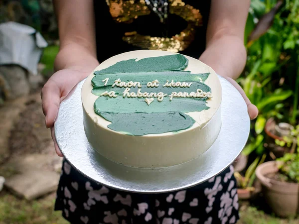 Girlfriend handing over a small cake with green icing and a heartwarming message. Tagalog words translated as 1 year and forever. Anniversary cake. Philippine dessert.