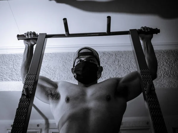 A man with no shirt and wearing a face mask and with an intense, determined look, prepares to do pull ups on a pull up bar.