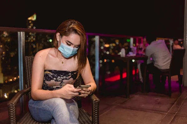 手術用マスクをした魅力的な若いアジア人女性は マニラ首都圏のマカティの屋上バーで凍えている間に携帯電話をチェックします — ストック写真