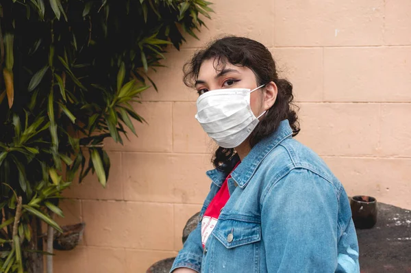 Kot ceketli, ameliyat maskesi takmış genç bir Filipinli kadın sarı tuğla duvarın ve yeşilliğin yanındaki taburede oturuyor..