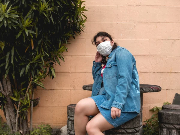 デニムジャケットを着た若いフィリピン人女性が 黄色いレンガの壁と緑の近くのコンクリートのスツールに座って手術用マスクをしている — ストック写真