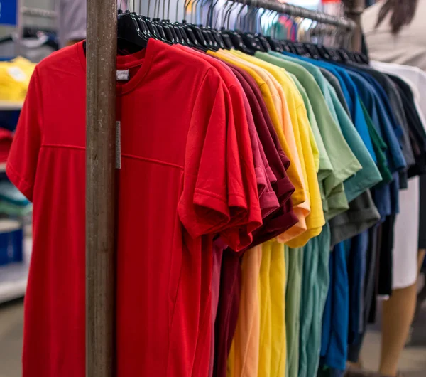 A selection of t shirts of a various colors displayed on a clothes rack at a department store.