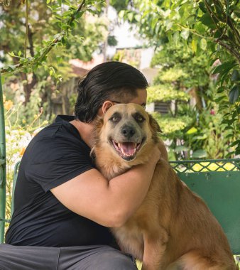 Siyah gömlekli bir adam, her ikisi de demir veranda salıncağında otururken dost köpeğine sarılıyor..
