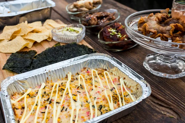 An assortment of food items on a wooden bench: Baked Sushi, Chicken wings, Chicharon Bulaklak and Baked Nachos. Outdoor party concept.