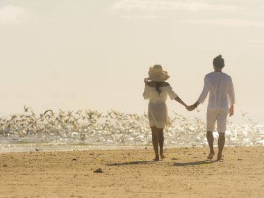 Beyazlar içinde el ele tutuşmuş bir çift sabah plajda yürürken arkadaki kırlangıç sürüsü uçup gidiyor. Samimi bir sabah sahili..