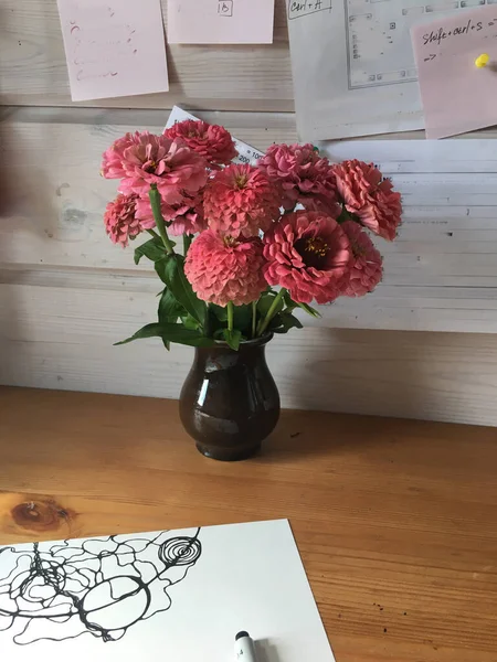 On the desktop of light wood is a sheet with a sketch of a drawing. there is an earthenware jug with a bouquet of pink zinnias. the wall is made of white wooden beams. There are notes pinned to it