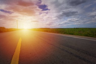 Çiftlik arazilerinin ve otlakların çevresindeki güzel asfalt yol gün batımında kırsal alanda, yoldaki sarı çizgi bulutlar ve mavi gökyüzü..