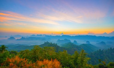 Gökyüzü mavi ve turuncu güneş ışığıyla güzel dağ sıraları gökyüzündeki bulutlar arasında, gündoğumu sırasında arka planda gökyüzü dağda sis, bereketli orman görüntüsü