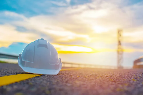 Close up of white safety helmet placed on the yellow solid line of road with sunset and blue sky background, Helmet in construction site after work during the sunset, Safety and Engineering Concept.