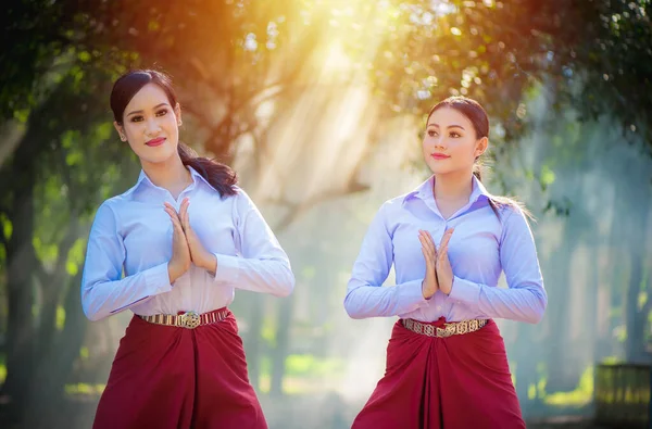 Two beautiful asian women friends with white shirt doing Thai gesture or begging (Wai). Welcome with a smile,The respect expresses the meaning to say hello. The sunshine with trees background.