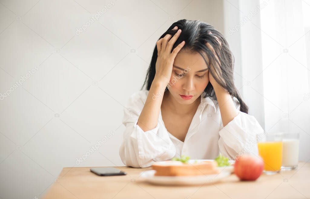 Beautiful Young female in white shirt uses his hands to hold the head to show stress with breakfast on table, Young woman suffering a headache, feeling sick and migraine from tired and health problem.
