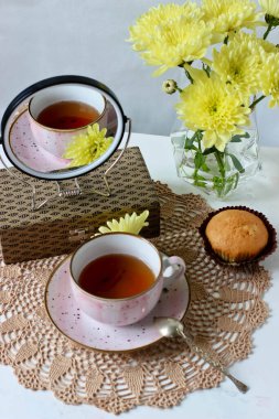 Aynada bir fincan çay ve örgü peçetede sarı kasımpatı olan bir vazo.