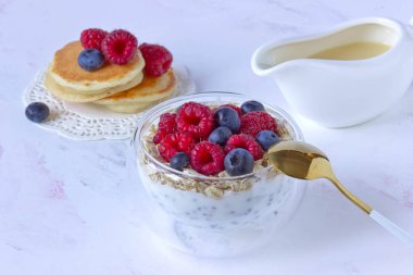 Şeffaf bir kasede yulaf ezmesi, ahududu ve yaban mersinli Chia pudingi soslu bir teknenin arka planında çift taban ve beyaz arka planda yoğunlaştırılmış süt ve krep.