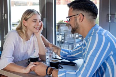 Genç romantik çift bir kafede kahvelerini yudumluyor, masada oturup gülümsüyor ve sohbet ediyorlar. Erkek arkadaşı nazikçe kız arkadaşına dokunuyor..