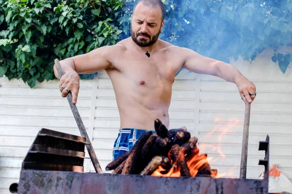 Barbekü yangın başlatma ustası. Genç kaslı adam kömür ızgarasını tutuşturmak için mısır koçanı kullanıyor.. 