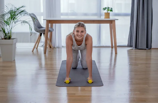 Beautiful active young woman work out at home. Half plank pose with dumbbells. Looking at camera. Healthy lifestyle and sports concept. Series of exercise poses.