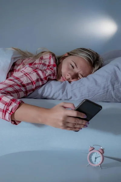 Beautiful young woman in pajamas sleeping in bed, turning off the alarm clock on a mobile phone, having a hard time to wake up.