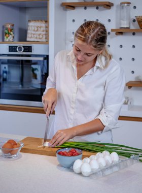 Modern bir mutfakta yemek pişiren güzel genç bir kadın. Taze soğan dilimlemek, yumurtalı kahvaltı hazırlamak. Evde yalnızım, tek kişilik yemek yapıyorum..