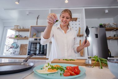 Modern donanımlı bir mutfakta yemek pişiren güzel genç bir kadın. Tavada kızarmış yumurtanın üzerine doğranmış taze soğan serpiyorum. Evde yalnızım. Seçici odak.