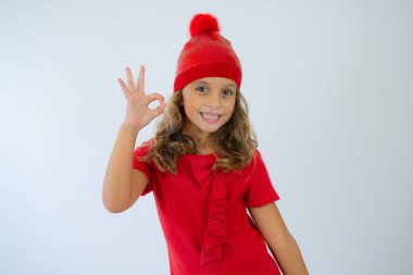 Stüdyoda beyaz arka planda örgü kırmızı şapka ve şirin kırmızı elbise giyen mutlu küçük kızın portresi. Moda sonbaharı kış sezonu indirimi konsepti. Noel gecesi ve doğum günü dileği diliyor.
