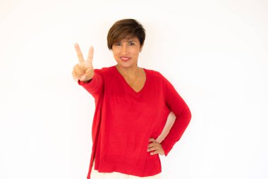 Güzel, esmer, kırmızı süveter giyen genç bir kadın izole edilmiş arka planda mutlu zafer jestleri yapıyor. Kameraya bakıp başarıyı gösteren ifade onaylanıyor.