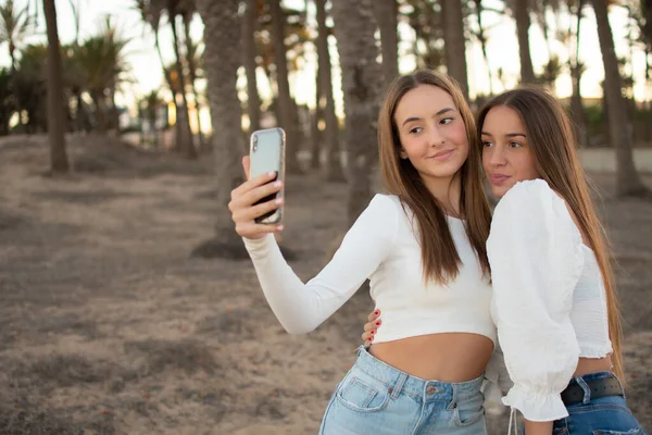 Güneşin doğuşunda arka planda sıcak bir ışıkla parkta selfie çeken iki mutlu kız arkadaş.