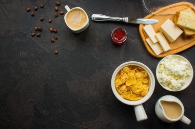 Mısır gevreği, kahve, süzme peynir, ekmek, tereyağı, yumurta, reçel ve diğer malzemeler için karanlık bir arka plan üzerinde kahvaltı. Kopya alanı ile üstten görünüm