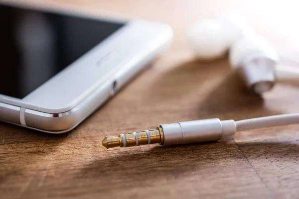 У вухах навушники і кабель лежать поруч з білим мобільним телефоном на дерев'яному столі — стокове фото