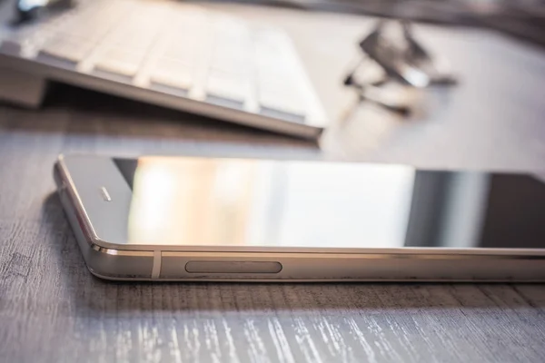 Smartphone mit Spiegelung, Brille und weißer Tastatur auf dem Tisch - Büroarbeitsplatzkonzept — Stockfoto