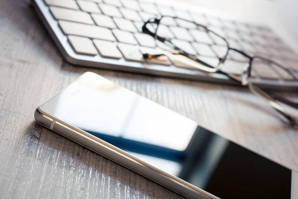 Teléfono móvil con reflexión, un par de gafas y un teclado blanco en una mesa - Concepto de lugar de trabajo de oficina —  Fotos de Stock