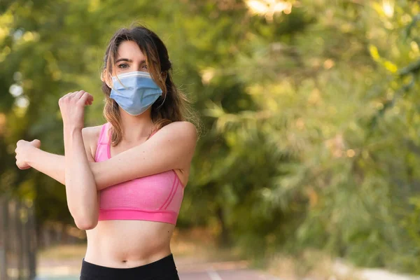 Covid virüsünden korunmak için maskeyle spor yaptıktan sonra kol kaslarını esneten bir kız.