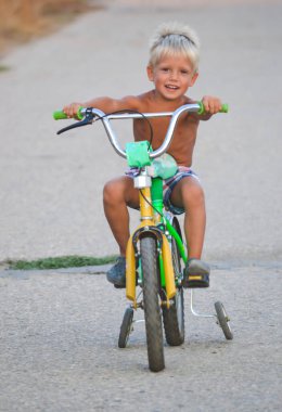 Küçük çocuk yaz günü yeşil bisiklete binmeyi öğreniyor. 4 tekerli bisikletli açık hava aktivitesinin mutluluğu, öğretim süreci olarak. Etkin genç çocukla asfalt arkaplan.