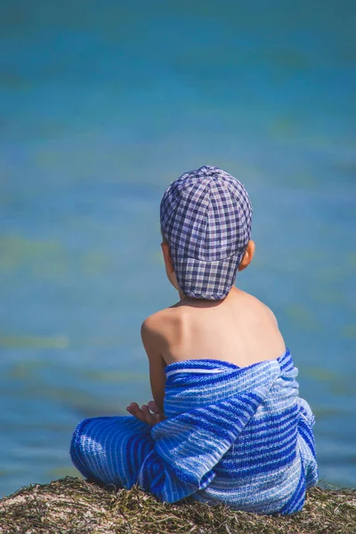 Small boy in hat and blue towel is sitting on the beach and looking on the sea. Blue color of water matching to towel color shade. Isolated relaxation on the beach. Alone tourism.