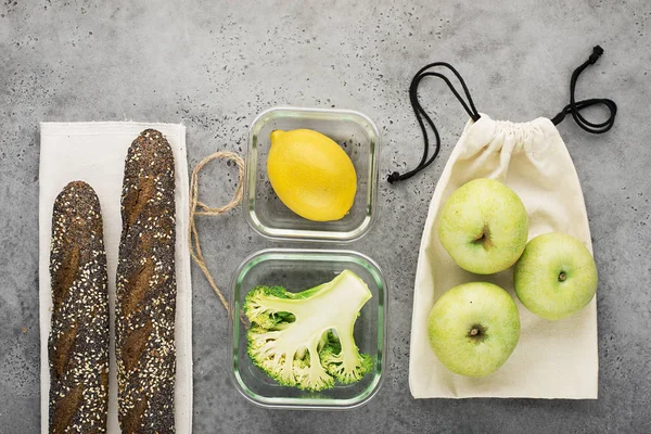 Eco bag pack with apples. Glass containers of broccoli, lemon, bread in a bread bag, wooden spoon and fork on a concrete gray background. Top view. Copy space.
