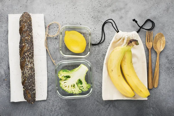 Eco bag pack with apples. Glass containers of broccoli, lemon, bread in a bread bag, wooden spoon and fork on a concrete gray background. Top view. Copy space.