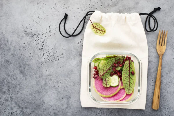 Watermelon radish cucumbers pomegranate rice sorrel salad in a glass container on eco bag. Snack with a wooden fork. Zero waste house. Top view. copy space.