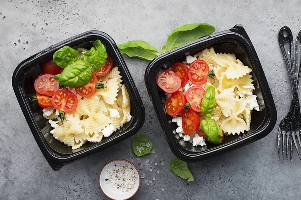 Meal prep. Black lunch boxes. Pasta bows, cherry tomatoes, young cheese, salt, pepper, basil. On a dark background Top view.