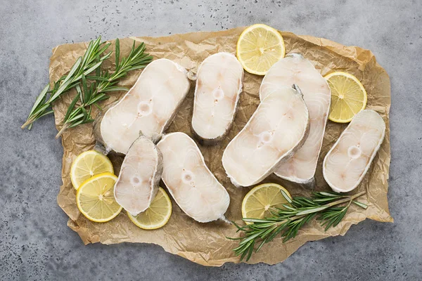 Filetes de bacalao blanco marino crudos sobre papel con ramitas de romero y rodajas de limón sobre un fondo gris claro. Vista superior . Imagen De Stock