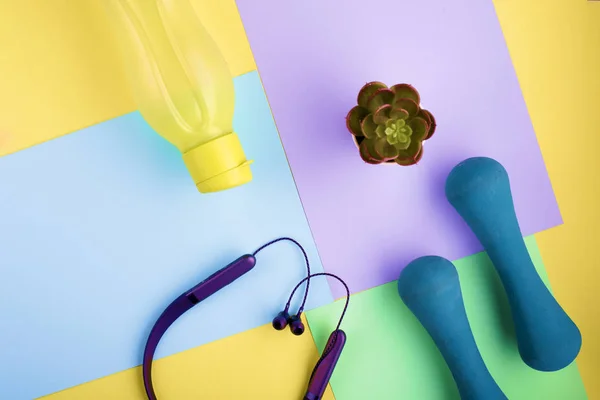 Moderne Kopfhörer, blaue Hanteln, gelbe Wasserflasche auf bunten Farbblöcken. Lifestyle-Konzept in Bewegung. hellen Kunstobjekten Hintergrund. horizontal, fototonisch, ultraviolett — Stockfoto