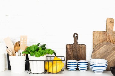 Mutfak masasında çatal bıçak, kaşık, spatula, taze fesleğen, kesme tahtası, taze sebzeler, basit bir duvarda limon. Yatay, boşluk,