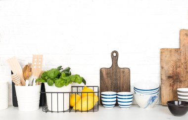 Mutfak masasında çatal bıçak, kaşık, spatula, taze fesleğen, kesme tahtası, taze sebzeler, basit bir duvarda limon. Yatay, boşluk,