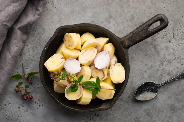 Нарезанный фиолетовый молодой сырой картофель перед выпечкой в кастрюле с луком, маслом и орегано. Вид сверху. Удобная домашняя еда. Плоский лежал, — стоковое фото