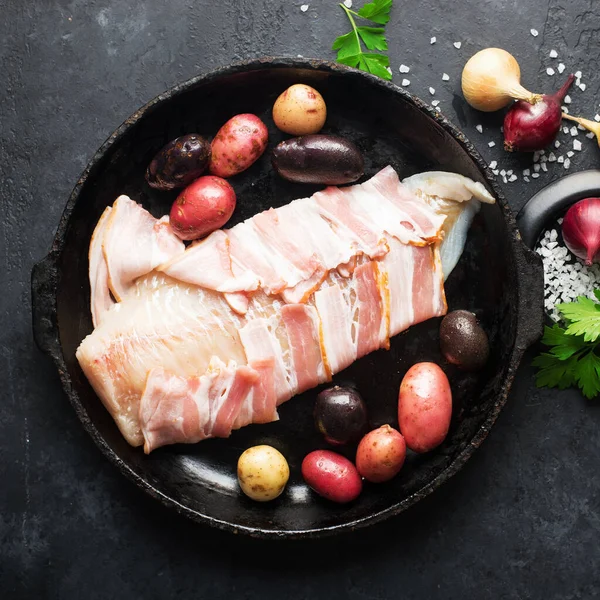 White cod fish wrapped in raw smoked bacon slices in a baking dish with new potatoes, spices and herbs. Top view. On a dark background Stock Image
