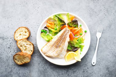 Kızarmış balık fileto, sulu marul, kapsika, limon, yeşil bezelye ve gri arka planda büyük beyaz bir tabakta. Sağlıklı dengeli yiyecekler. Üst görünüm