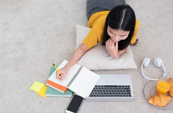 最热门看法亚洲妇女躺在地毯上 写在笔记本上 并学习在笔记本电脑与放松情绪在家中 工作在家里的概念 — 图库照片