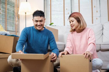 Mutlu Asyalı çift oturma odasındaki kanepeye taşınırken karton kutuları açıyor. Yeni ev için bavulları açıyorlar.